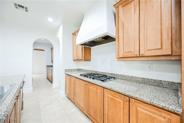 kitchen with custom exhaust hood, light tile patterned floors, light stone countertops, stainless steel gas cooktop, and sink