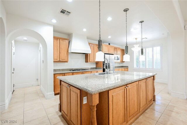 kitchen with custom exhaust hood, a kitchen island with sink, sink, pendant lighting, and light stone counters