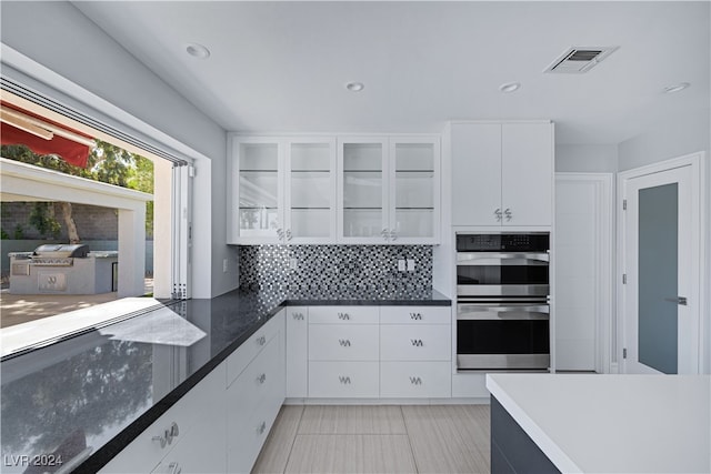 kitchen with stainless steel double oven, white cabinetry, light tile patterned floors, and backsplash