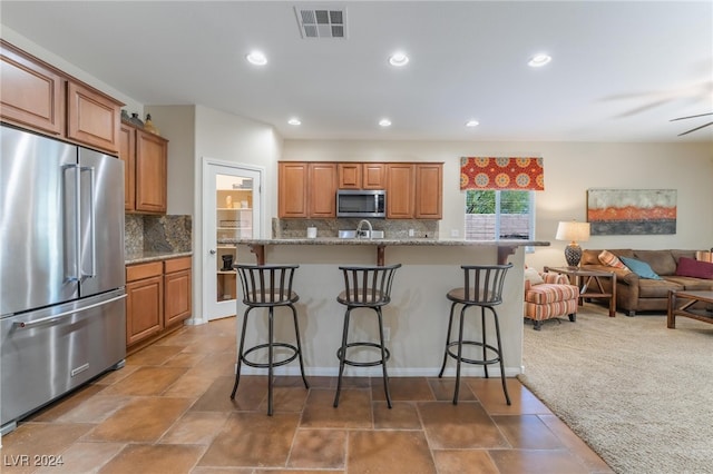 kitchen featuring appliances with stainless steel finishes, light stone counters, an island with sink, a breakfast bar, and backsplash