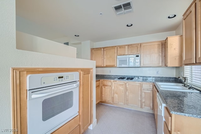 kitchen featuring dark stone counters, white appliances, light brown cabinetry, and sink