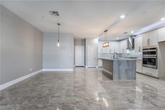 kitchen featuring wall chimney exhaust hood, stainless steel double oven, a center island with sink, white cabinets, and hanging light fixtures