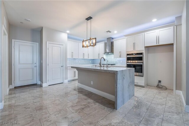 kitchen with a center island with sink, white cabinets, hanging light fixtures, wall chimney exhaust hood, and stainless steel double oven