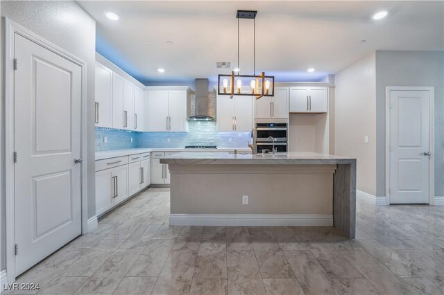 kitchen featuring double oven, wall chimney range hood, decorative light fixtures, a center island with sink, and white cabinetry