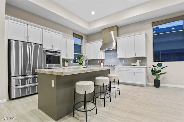 kitchen with white cabinetry, appliances with stainless steel finishes, wall chimney range hood, an island with sink, and a breakfast bar