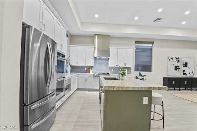 kitchen with appliances with stainless steel finishes, a kitchen breakfast bar, white cabinets, wall chimney exhaust hood, and a kitchen island with sink