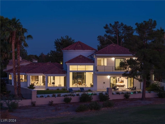 back of house at night featuring a balcony, a patio area, a fenced front yard, and stucco siding