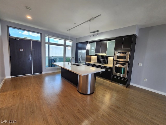 kitchen with an island with sink, sink, wood-type flooring, decorative light fixtures, and appliances with stainless steel finishes