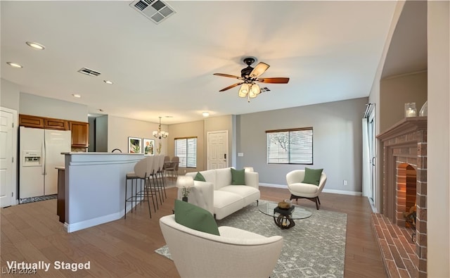 living room featuring light wood-type flooring, ceiling fan with notable chandelier, and a brick fireplace