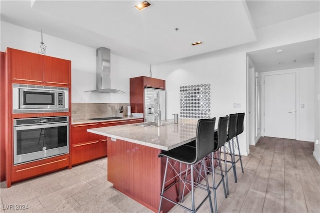 kitchen with wall chimney exhaust hood, decorative backsplash, an island with sink, a kitchen bar, and stainless steel appliances