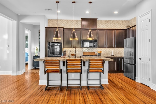 kitchen with a center island with sink, hanging light fixtures, light stone countertops, black appliances, and light hardwood / wood-style floors