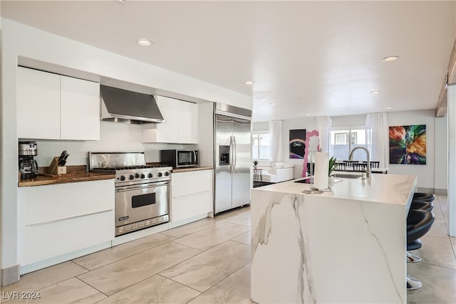 kitchen featuring a kitchen island with sink, white cabinets, wall chimney range hood, sink, and premium appliances