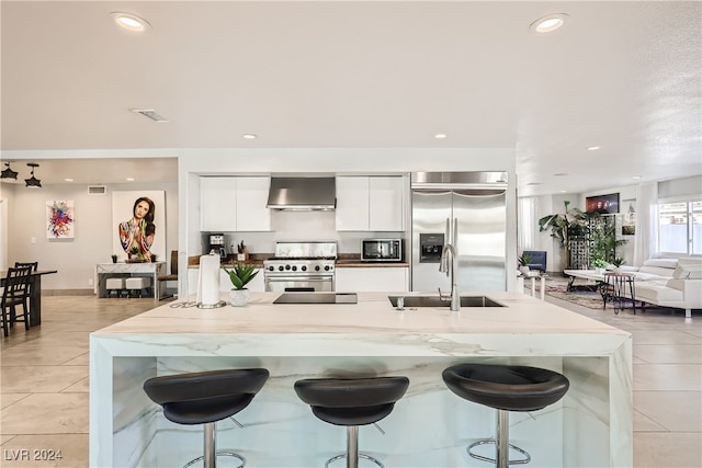 kitchen featuring a kitchen bar, high end appliances, sink, wall chimney range hood, and white cabinetry