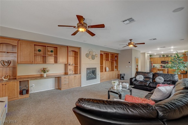 living room featuring ceiling fan, built in desk, carpet flooring, and a tiled fireplace