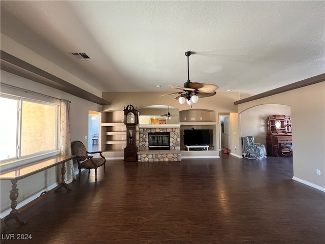 living room featuring dark wood-type flooring, a stone fireplace, ceiling fan, a textured ceiling, and built in shelves