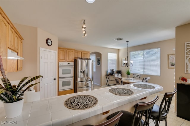 kitchen featuring stainless steel fridge, a chandelier, tile counters, white double oven, and light brown cabinetry