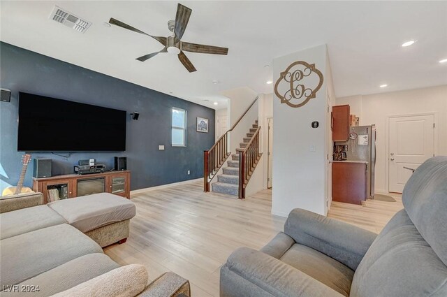living room featuring ceiling fan, light wood-type flooring, an accent wall, visible vents, and stairway