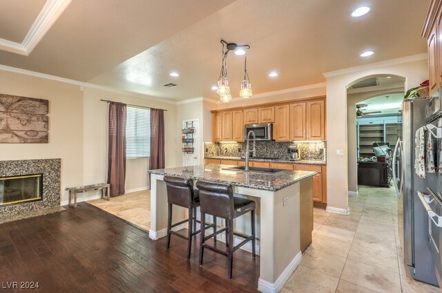 kitchen featuring arched walkways, a center island with sink, stainless steel microwave, a high end fireplace, and dark stone counters