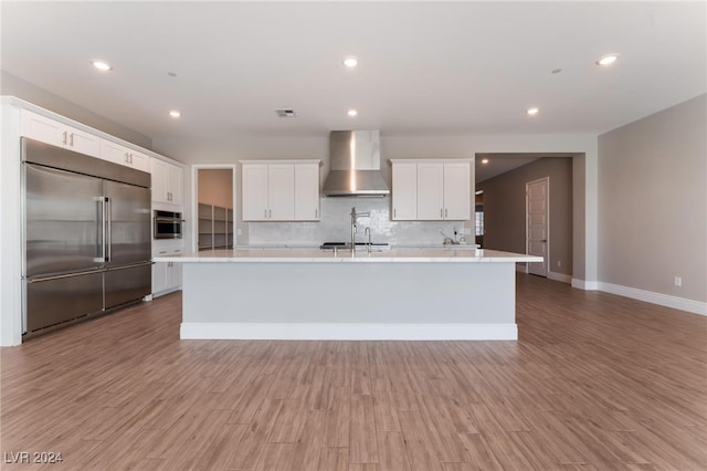 kitchen with appliances with stainless steel finishes, a kitchen island with sink, white cabinets, decorative backsplash, and wall chimney exhaust hood