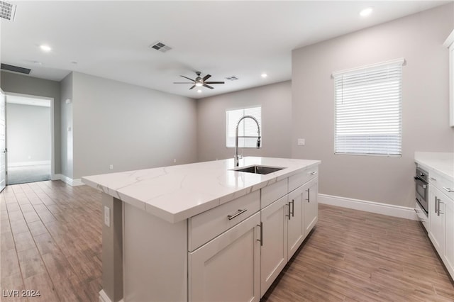 kitchen with sink, white cabinetry, stainless steel oven, light stone counters, and an island with sink