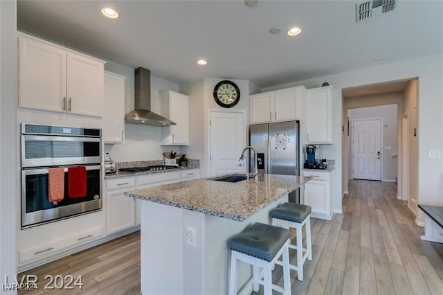 kitchen with sink, a center island with sink, wall chimney exhaust hood, white cabinetry, and appliances with stainless steel finishes