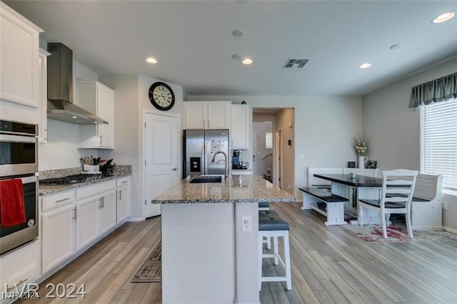 kitchen with white cabinets, wall chimney range hood, light hardwood / wood-style flooring, appliances with stainless steel finishes, and a center island with sink