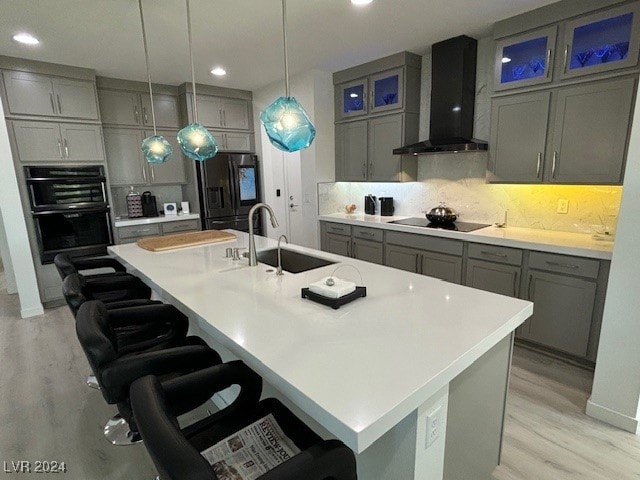 kitchen with wall chimney range hood, black appliances, sink, and a kitchen island with sink