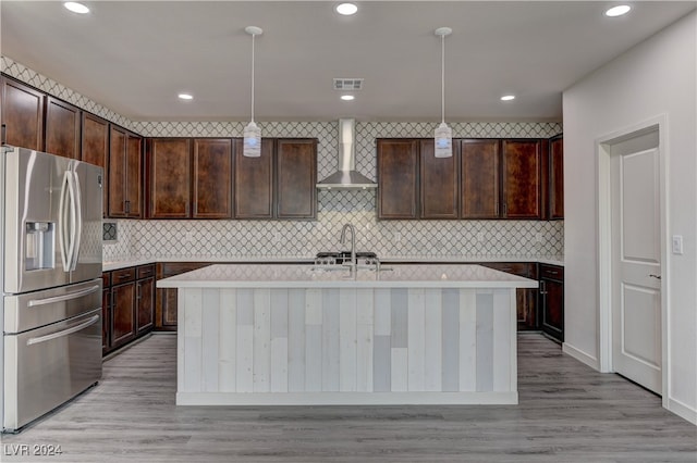 kitchen featuring wall chimney range hood, decorative light fixtures, stainless steel fridge, and a kitchen island with sink