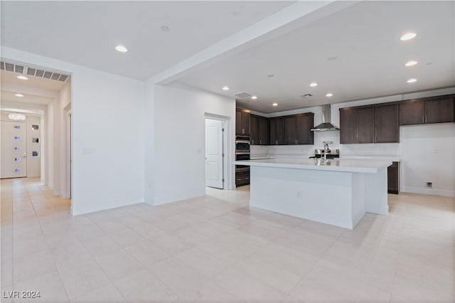 kitchen with double oven, sink, wall chimney exhaust hood, an island with sink, and dark brown cabinets