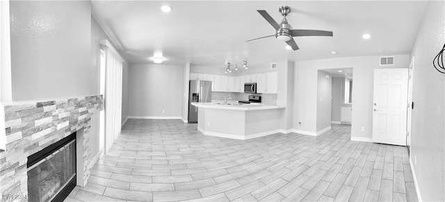 kitchen with a stone fireplace, kitchen peninsula, stainless steel appliances, white cabinetry, and ceiling fan