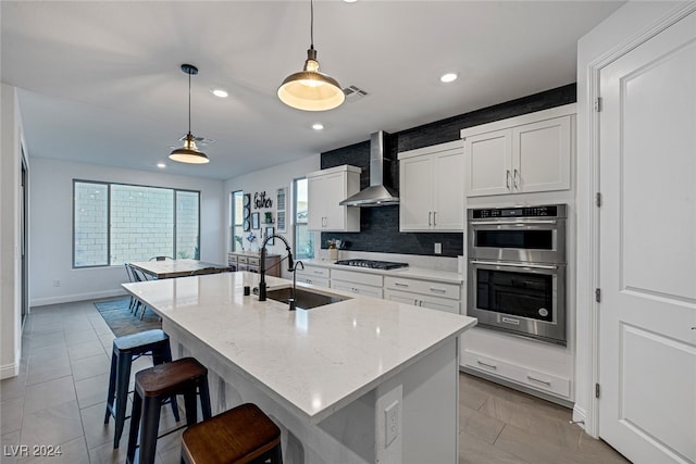 kitchen featuring wall chimney range hood, appliances with stainless steel finishes, a center island with sink, and hanging light fixtures