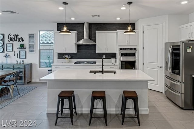 kitchen featuring wall chimney range hood, stainless steel appliances, a center island with sink, sink, and decorative light fixtures