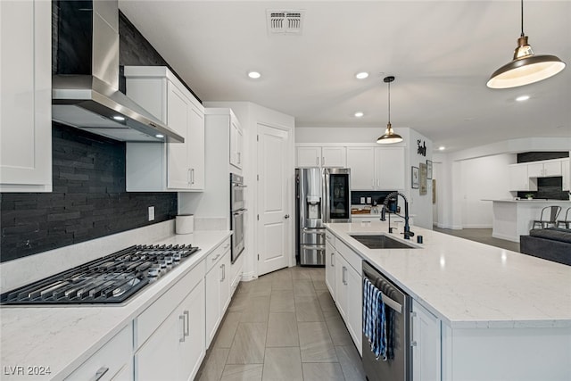 kitchen featuring wall chimney range hood, a center island with sink, pendant lighting, white cabinetry, and appliances with stainless steel finishes