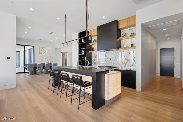 kitchen featuring a kitchen bar, an island with sink, wall chimney range hood, light hardwood / wood-style floors, and backsplash