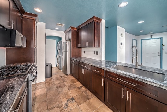 kitchen featuring dark brown cabinets, dark stone countertops, stacked washer and dryer, sink, and stainless steel appliances