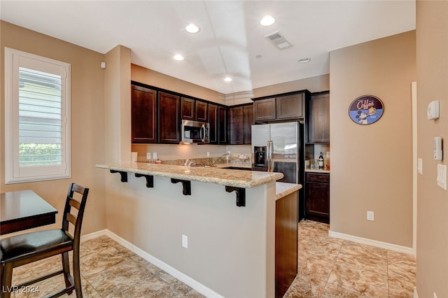 kitchen featuring kitchen peninsula, dark brown cabinets, a breakfast bar area, appliances with stainless steel finishes, and light stone countertops