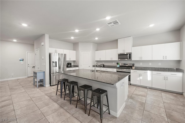 kitchen featuring a breakfast bar area, appliances with stainless steel finishes, white cabinetry, a kitchen island with sink, and dark stone counters