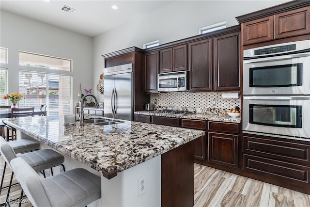 kitchen with an island with sink, backsplash, stainless steel appliances, light stone counters, and a breakfast bar area