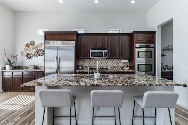 kitchen with appliances with stainless steel finishes, dark brown cabinetry, light wood-type flooring, and a kitchen island with sink