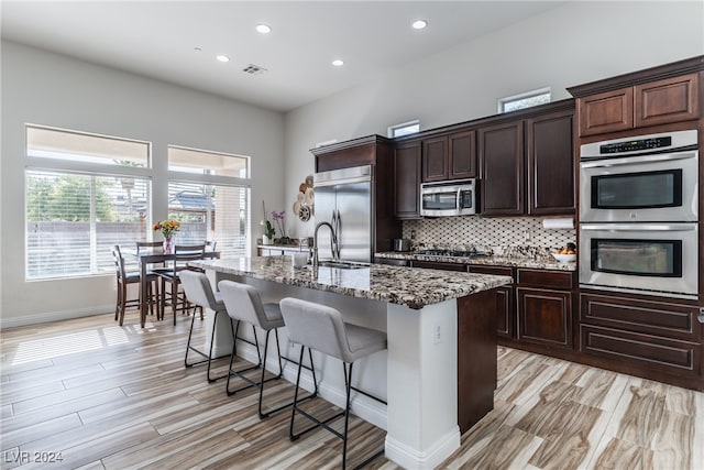 kitchen featuring an island with sink, backsplash, a breakfast bar, appliances with stainless steel finishes, and light stone counters