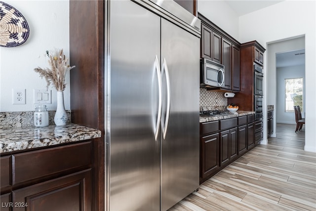 kitchen with tasteful backsplash, light stone countertops, dark brown cabinets, stainless steel appliances, and light hardwood / wood-style flooring