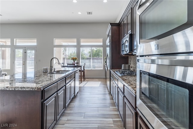 kitchen with stainless steel appliances, a center island with sink, sink, light wood-type flooring, and light stone counters