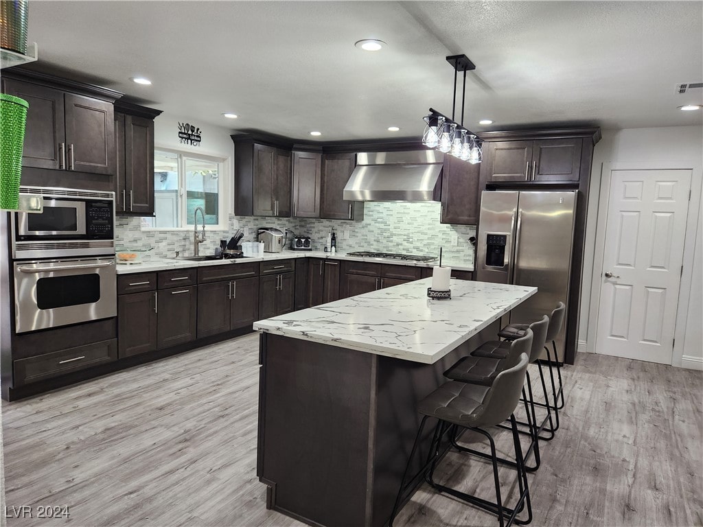 kitchen with stainless steel appliances, wall chimney exhaust hood, sink, pendant lighting, and light wood-type flooring