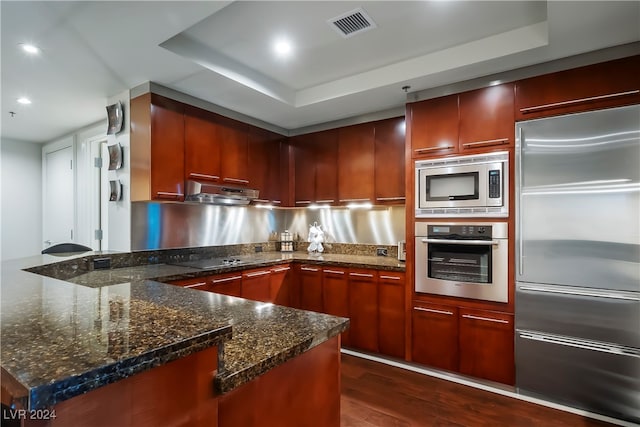 kitchen with built in appliances, a tray ceiling, dark hardwood / wood-style flooring, and dark stone counters