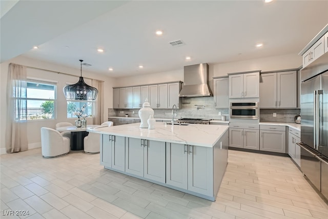 kitchen with wall chimney range hood, a center island with sink, appliances with stainless steel finishes, light stone countertops, and pendant lighting