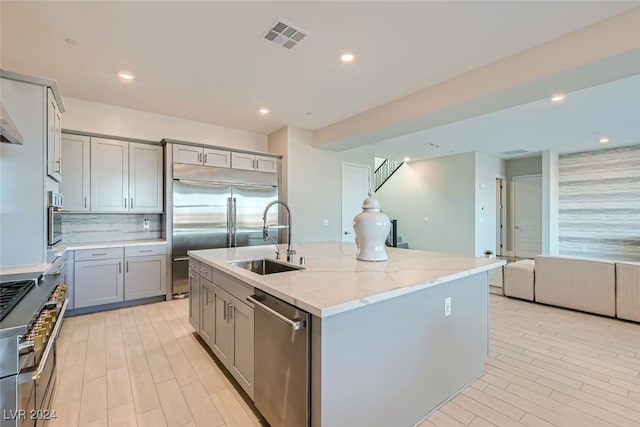 kitchen with gray cabinetry, high quality appliances, light stone counters, and an island with sink