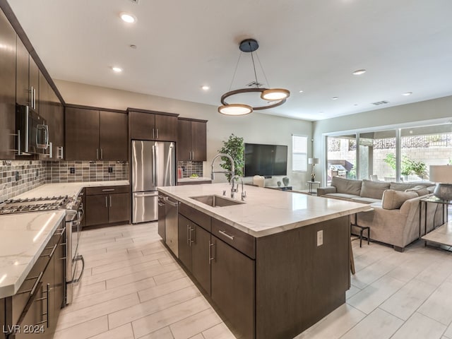 kitchen with an island with sink, backsplash, appliances with stainless steel finishes, dark brown cabinets, and sink