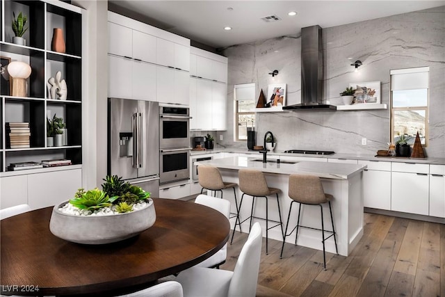 kitchen featuring appliances with stainless steel finishes, wall chimney exhaust hood, a kitchen island with sink, sink, and white cabinetry