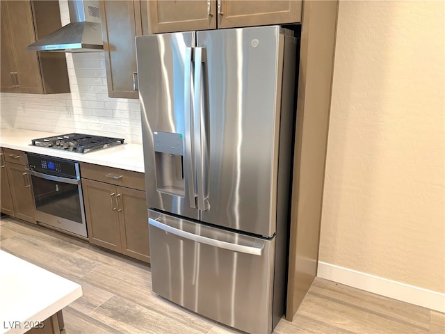 kitchen featuring light wood-type flooring, wall chimney range hood, backsplash, and appliances with stainless steel finishes