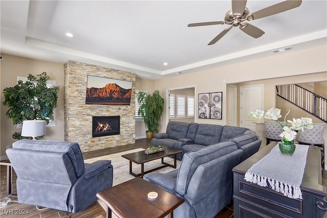 living room with dark wood-type flooring, ceiling fan, a tray ceiling, and a stone fireplace